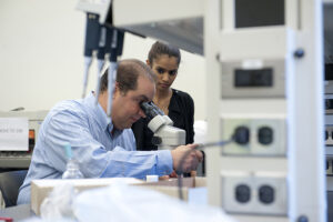 Dr. Fabrizio looks at a microscope and a student watches. 