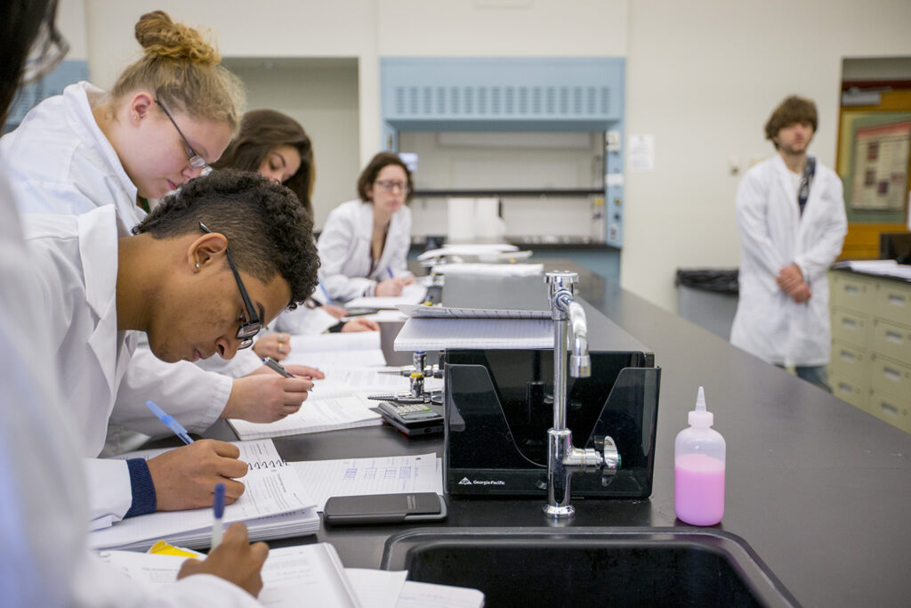 Four students write in notebooks, with one students off to the right frame of the image, in a science lab with black countertops and a sink in the front.