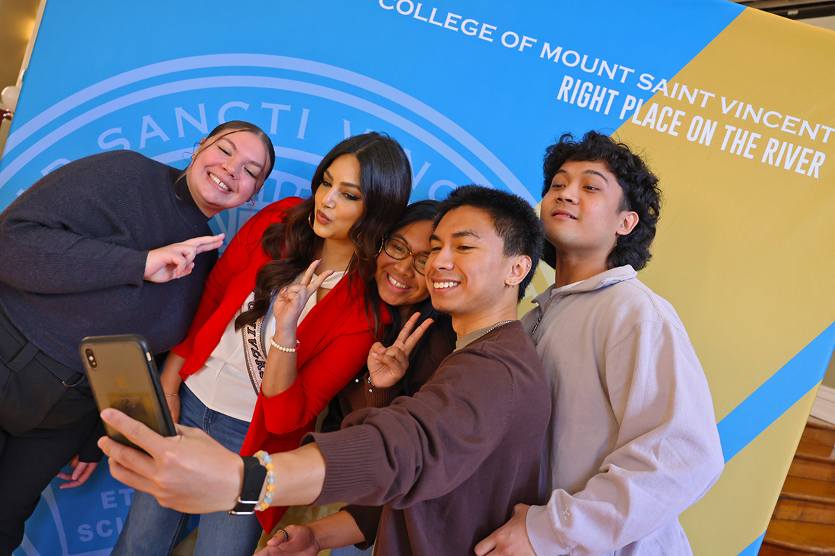 Miss Universe 2021 Harnaaz Sandhu poses in front of a blue and gold backdrop with a group of Mount Saint Vincent students to take a selfie