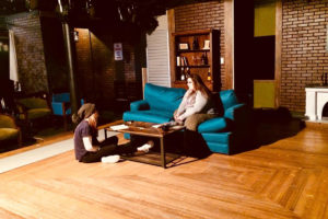 Two student actors talk while one is sitting on a couch and one is sitting on the floor in Stop Kiss at CMSV.