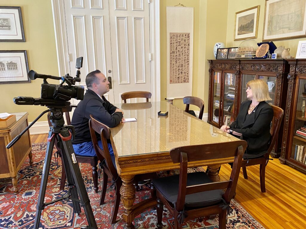 News 12 The Bronx reporter Dan Serafin interviews President Susan Burns at a table in an office with a camera filming them