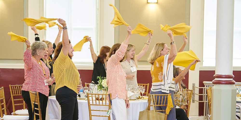 Alumnae from the College of Mount Saint Vincent celebrate at the 2021 luncheon; women at a dining table in a well-lit space raise their arms up in excitement and wave yellow napkins in the air