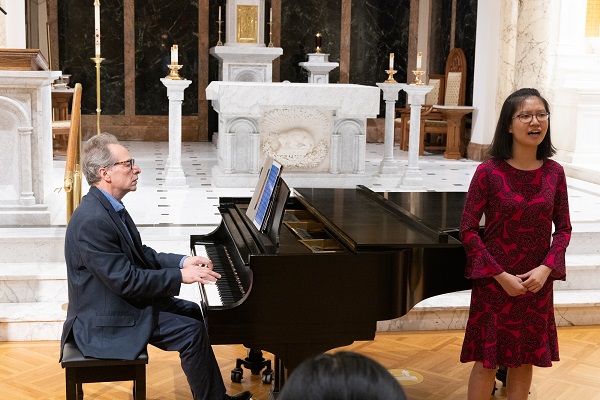 A pianist plays a piano as a female student in a red dress sings.