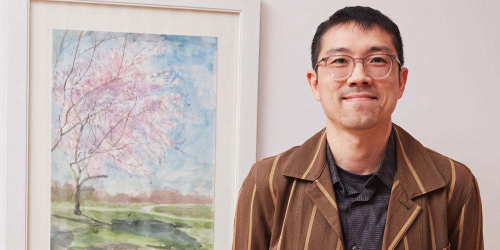 Photo of Professor Chemin Hsiao next to his watercolor painting in a white frame.