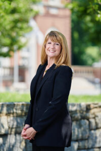 President Susan R. Burns, PhD, stands in front of a stone wall on the Mount Saint Vincent campus, with greenery in the background, wearing a black blazer with her hands clasped 