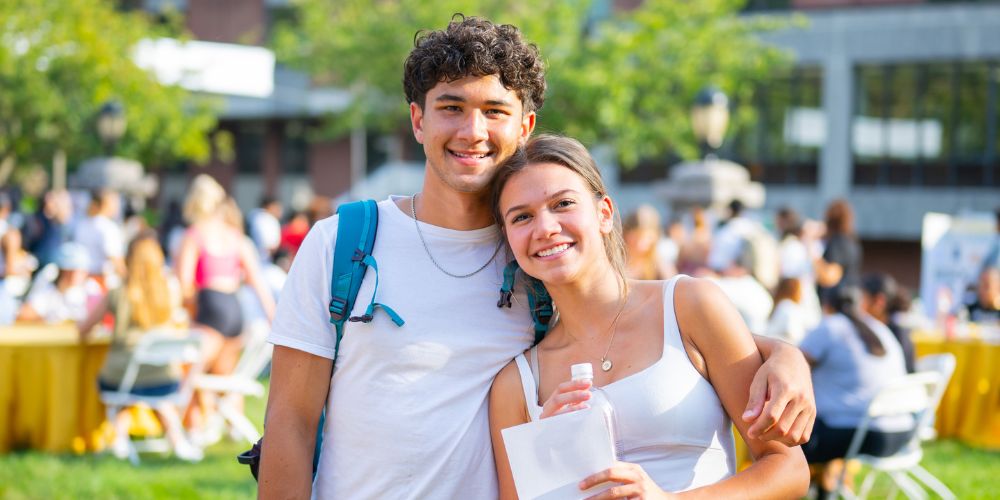A male student smiles and stands next to a smiling female student at an outside campus life activities fair
