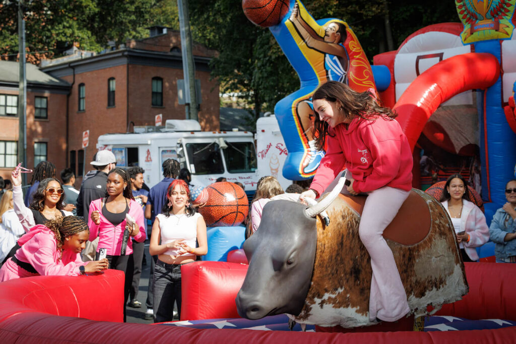Students participate in a University of Mount Saint Vincent homecoming event, with a student riding a mechanical bull surrounded by other students and colorful inflatable games.